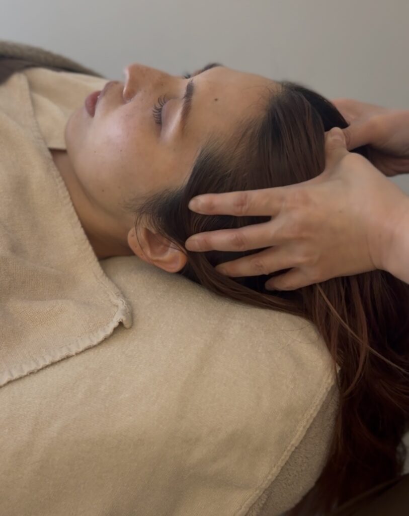 仰向けで横たわる女性の頭に手を添え、やさしく頭皮マッサージを行っている施術シーン。力が抜けてリラックスした表情から、安心して身をゆだねている様子が伝わる。